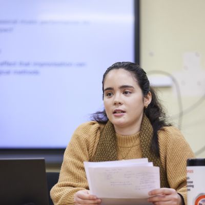 Doctoral student in a yellow jumper holding notes while sat at a desk